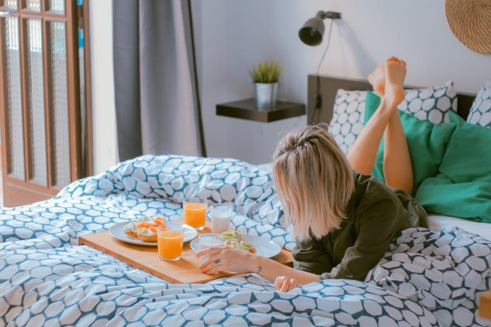 Woman relaxing on her bed while eating from a food tray.